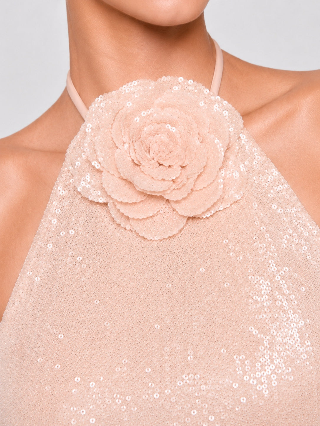 Close-up of a model wearing a pale blush pink halter top with a large layered fabric flower applique on the chest and scattered sequins across the fabric.