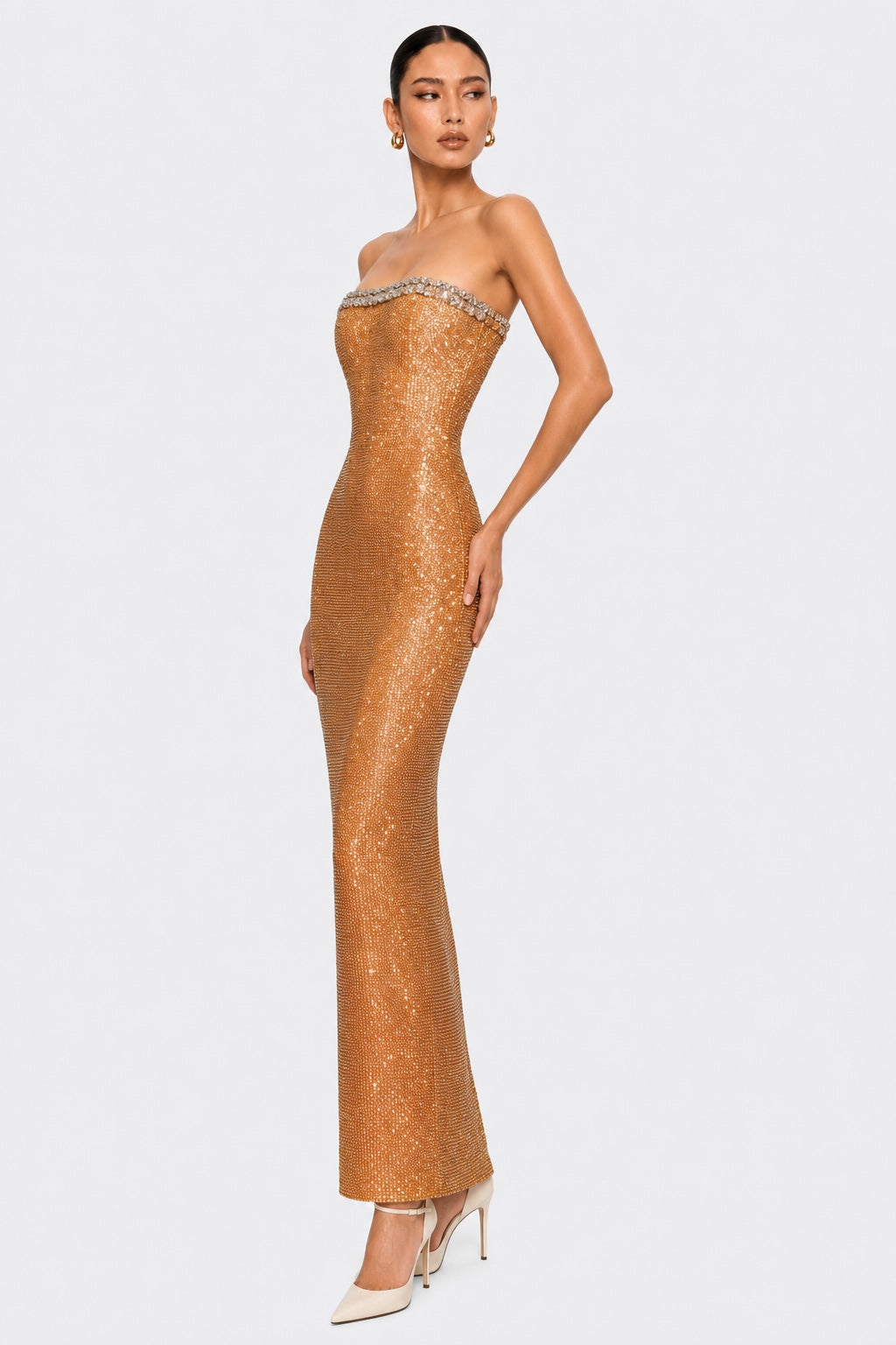 A woman modeling a strapless, floor-length metallic gold sequin gown with a crystal-embellished sweetheart neckline, paired with white pointed-toe heels, against a light gray studio background.