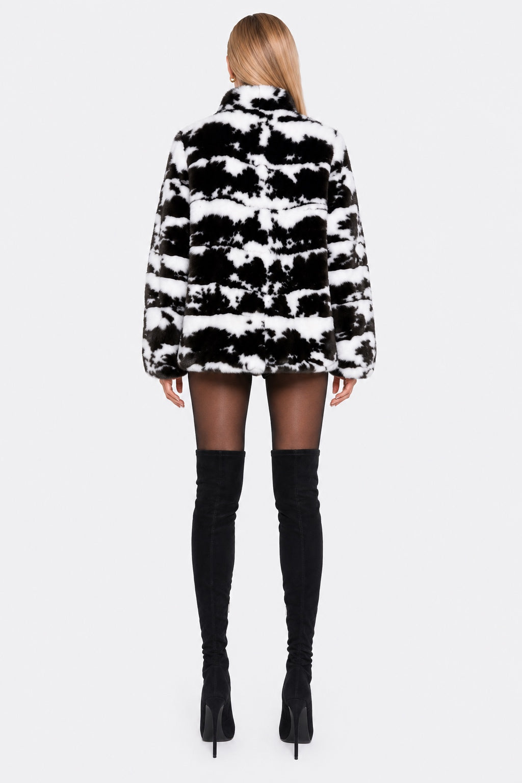 A woman shown from the back wearing a black and white patterned faux fur jacket, sheer black tights, and black over-the-knee boots against a light gray studio background.