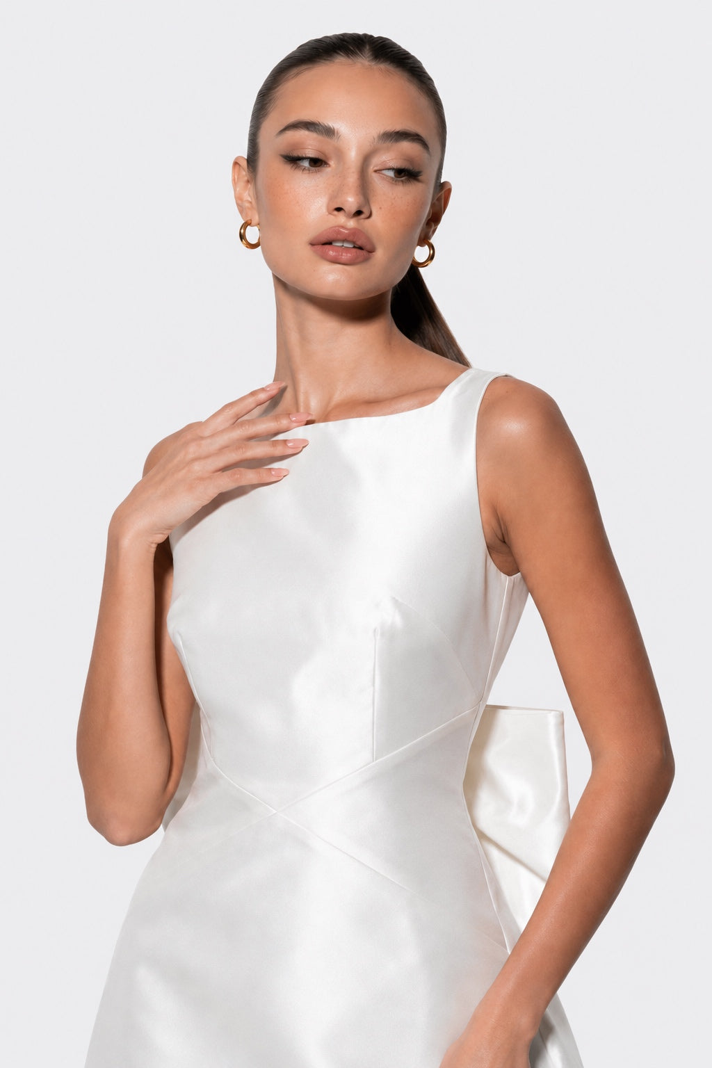 A woman modeling a sleeveless white satin dress with a scoop neckline and structured bodice, photographed against a light gray studio background.
