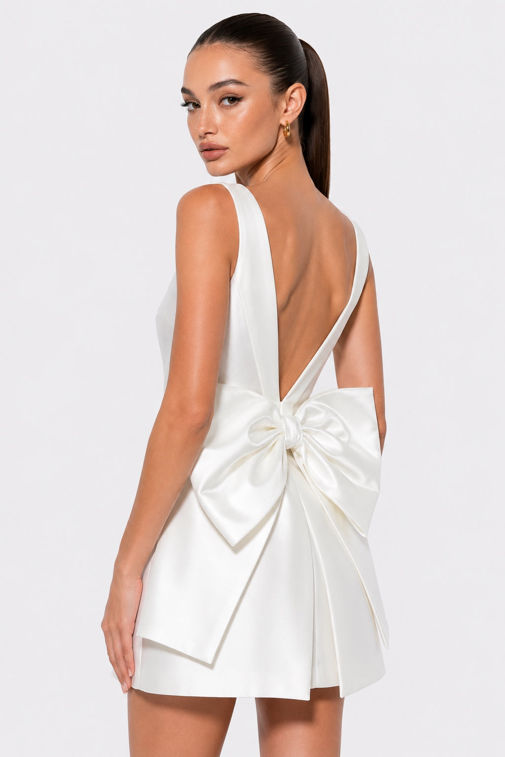 A woman modeling a white satin mini dress from the back, featuring a large bow detail at the back of the open neckline, set against a plain light gray studio background.