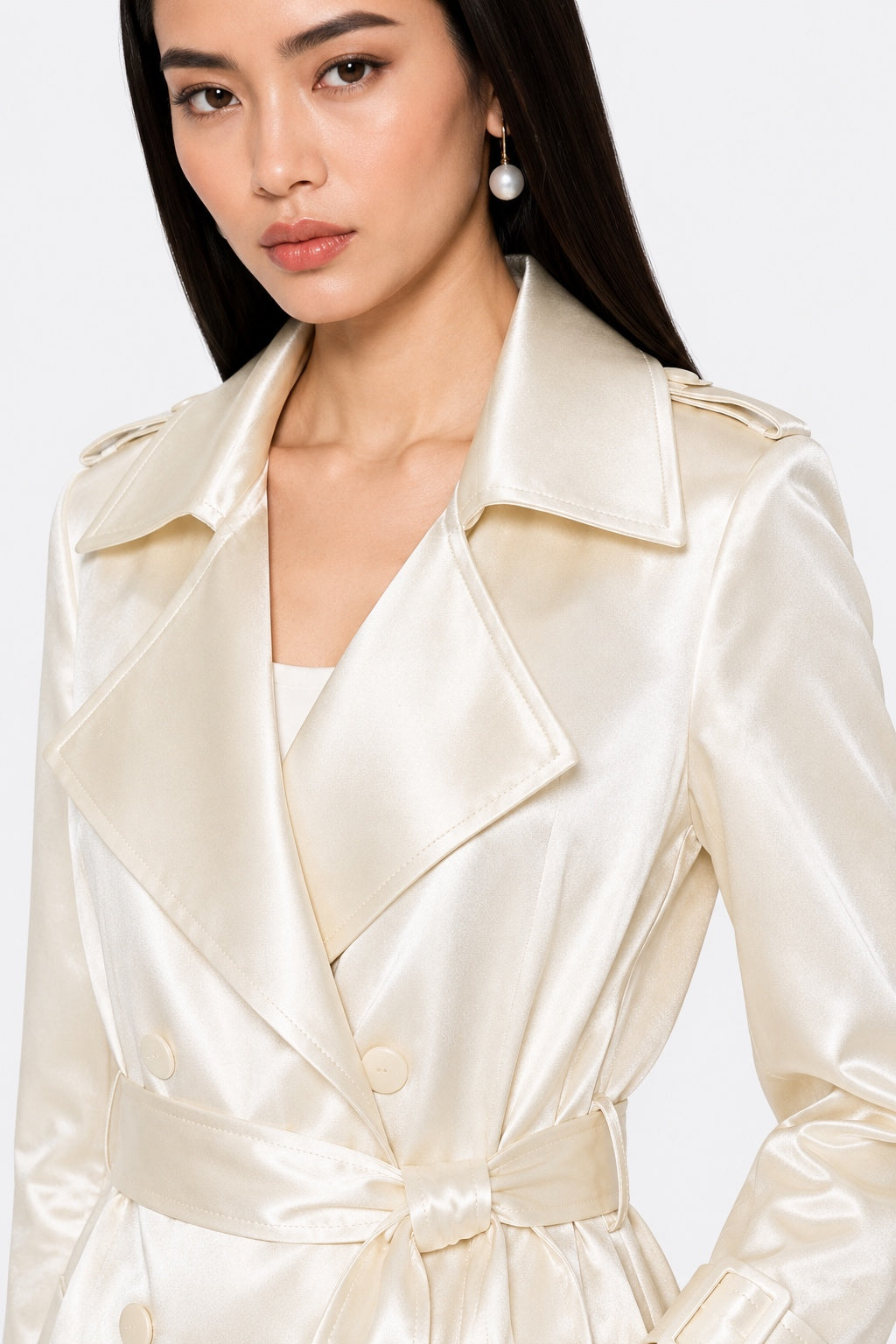 A close-up of a model wearing a cream-colored satin trench coat with a matching waist tie belt, set against a plain light gray background.