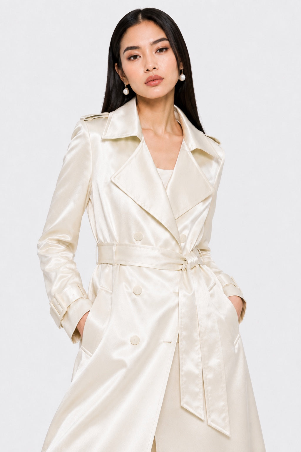 A woman modeling a cream-colored satin trench coat with a matching waist tie belt, set against a plain light gray studio background.