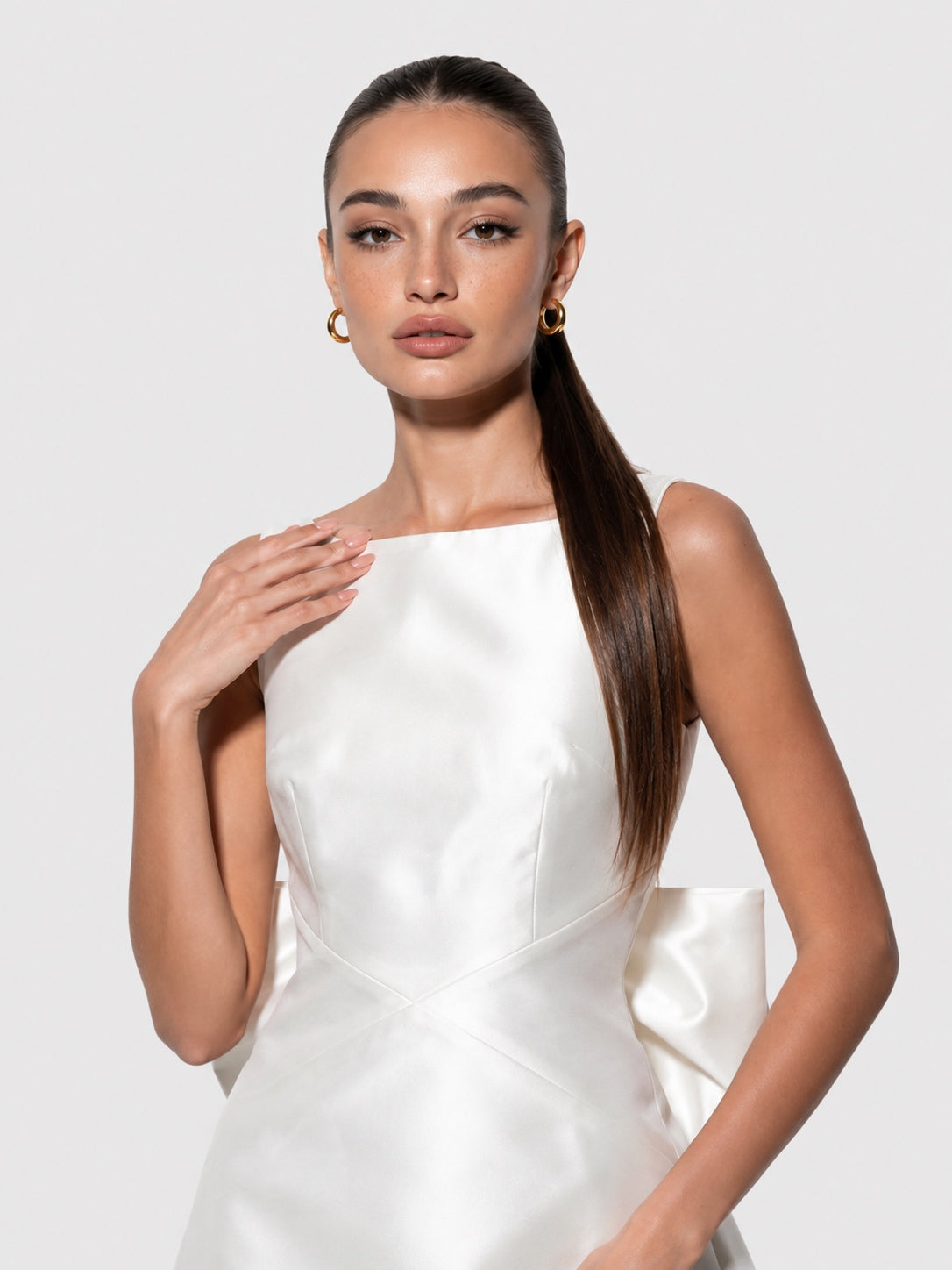 A woman with her hair in a ponytail wears a white satin sleeveless dress with structured shoulders and gold hoop earrings, posed against a light gray studio background.