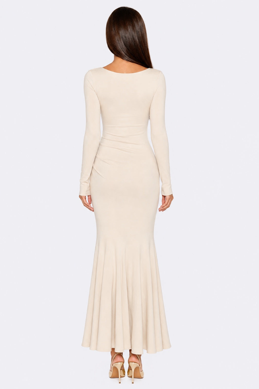 A woman viewed from the back wearing a fitted long-sleeve maxi dress in a warm cream or ivory tone, paired with beige high heels, against a plain light gray studio background.