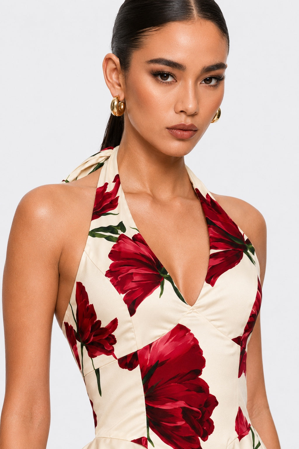 A close-up of a model wearing a halter-neck dress with a deep V-neckline, featuring a cream base with a bold red floral print and green leaves, set against a plain light gray background.