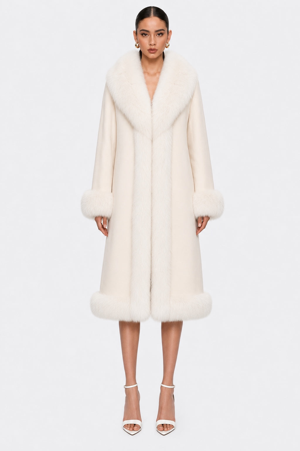 A model wearing a long, plush cream-colored natural fur coat with a wide collar, cuffs, and hem, paired with white high-heeled sandals, against a plain light gray background.