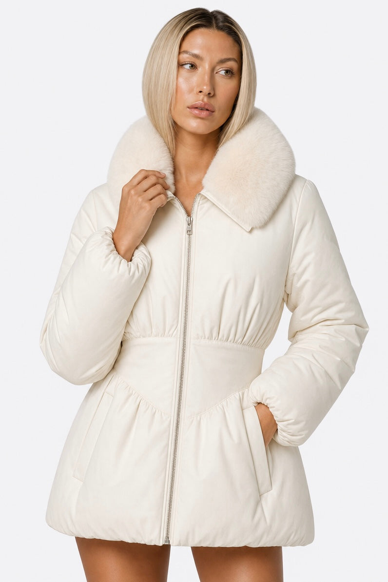 A woman wearing a cream-colored puffer jacket with a large fluffy faux-fur collar, photographed against a plain light gray studio background.