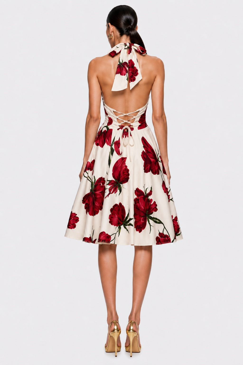 A woman viewed from the back wearing a sleeveless halter-neck midi dress with an open back and lace-up detail, featuring a red floral print on a cream background. She is wearing gold high heels against a light gray studio background.