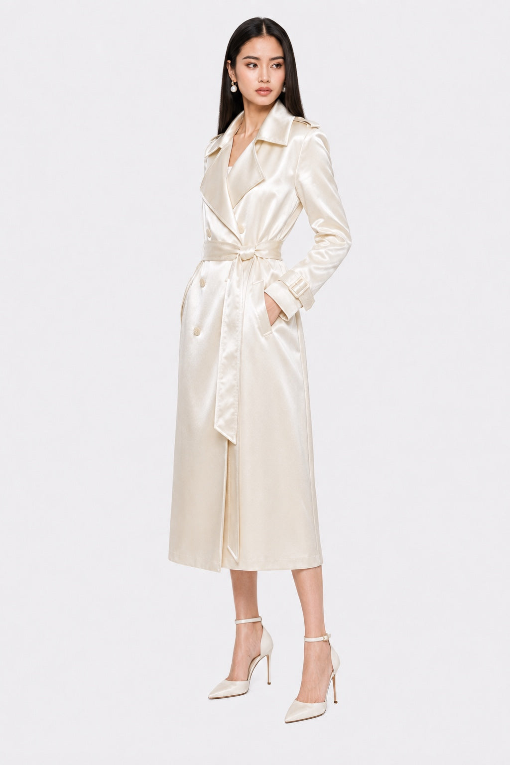 A woman modeling a long cream satin trench coat with a matching waist tie, paired with matching pointed-toe heels, against a light gray studio background.