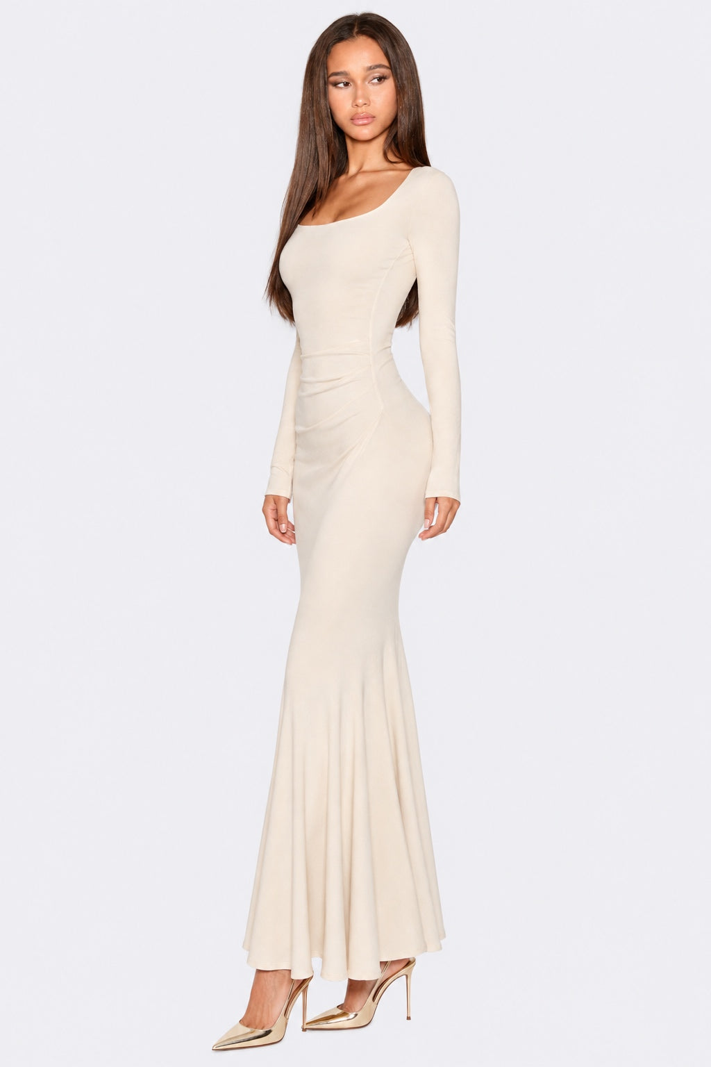 A woman modeling a long-sleeve, off-the-shoulder, fitted maxi dress in a warm cream or ivory shade, paired with metallic gold high heels, against a light gray studio background.