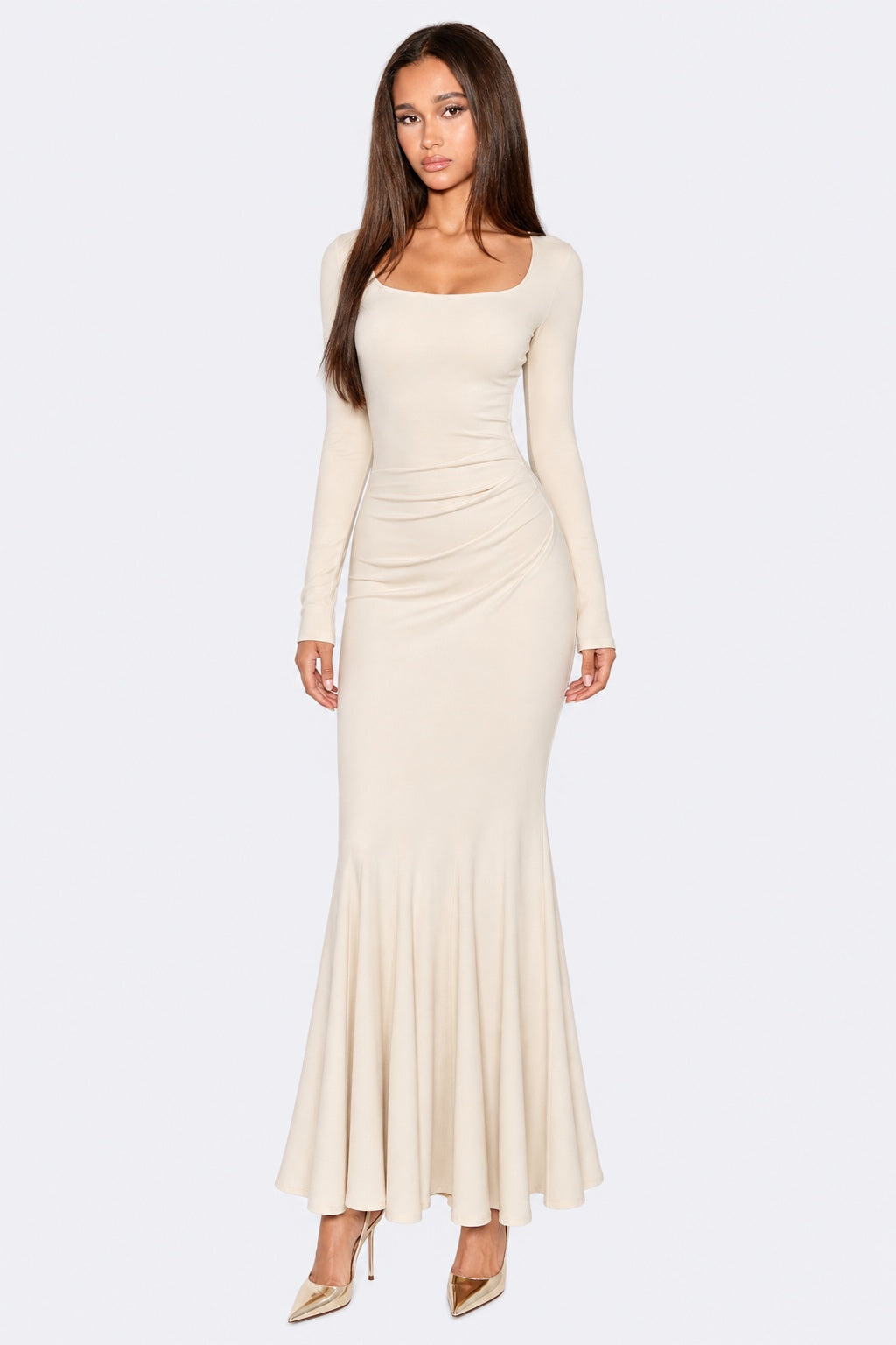 A woman modeling a long-sleeve, square-neck maxi dress in a light cream or off-white color, styled with metallic beige high heels against a light gray studio background.