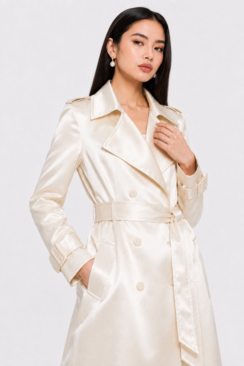 A woman modeling a cream satin trench coat with a matching waist belt against a light gray studio background.