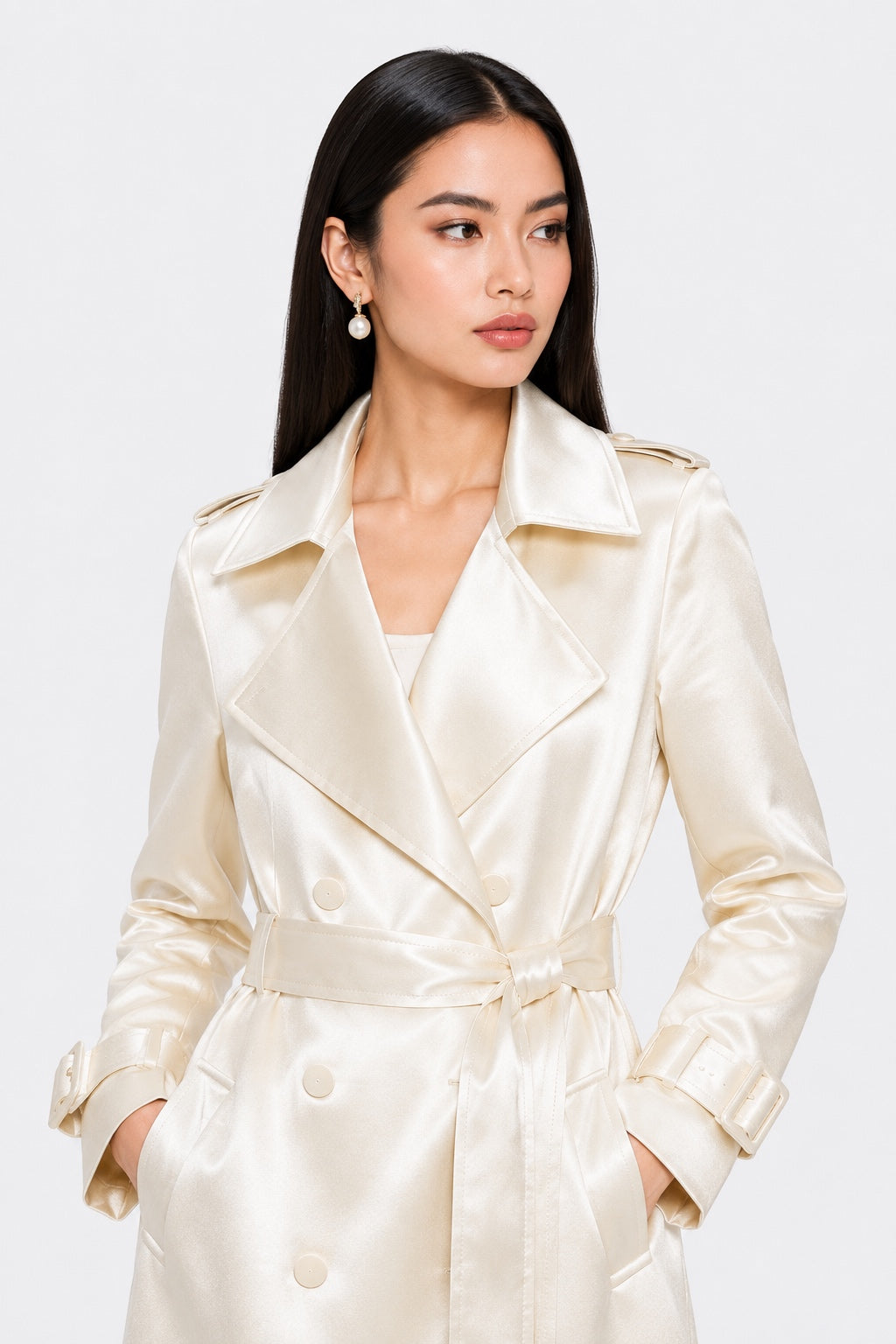 A woman modeling a cream-colored satin trench coat with a matching waist tie belt, set against a plain light gray studio background.