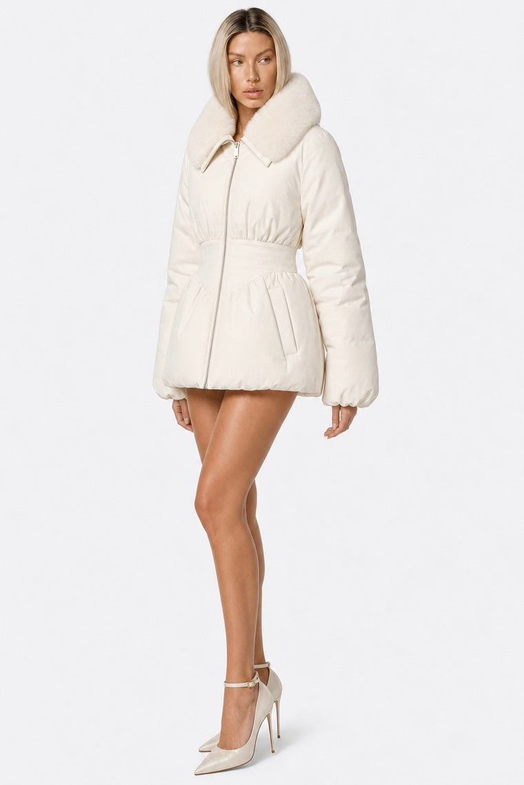 A model wearing a cream-colored puffer jacket with a large faux-fur collar and a matching belt, paired with cream high heels, against a plain white studio background.