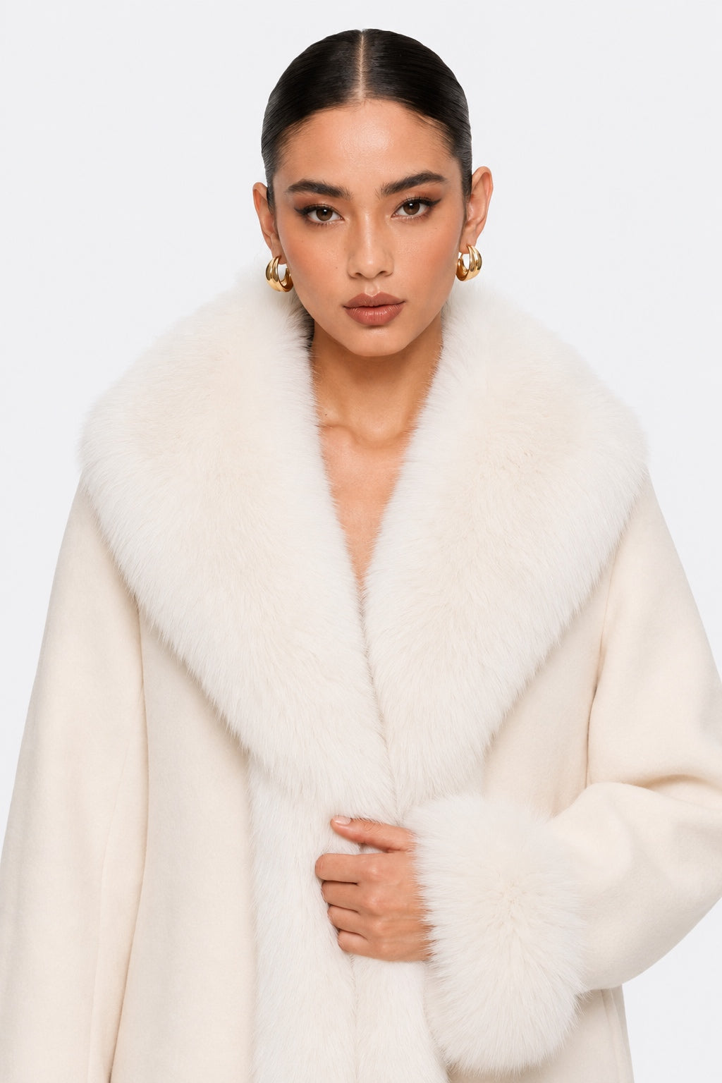 A model wearing a voluminous cream natural fur coat with a wide collar and matching cuffs, styled with gold hoop earrings against a light gray studio background.