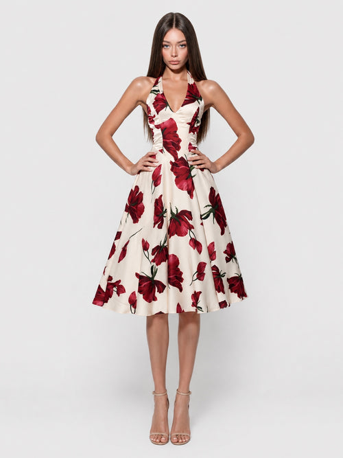 A woman modeling a sleeveless halter-neck midi dress with a red floral print on a cream background, paired with nude high-heeled sandals, against a light gray studio background.