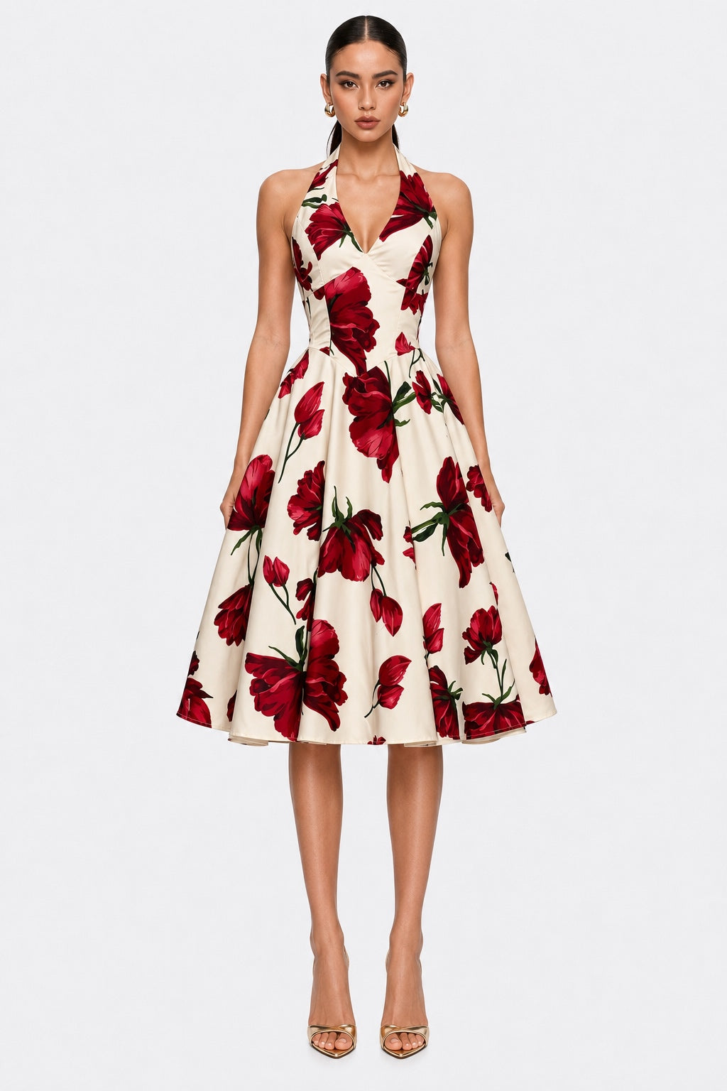 A woman modeling a sleeveless halter-neck midi dress with a red floral print on a cream background, paired with nude high heels, against a light gray studio background.
