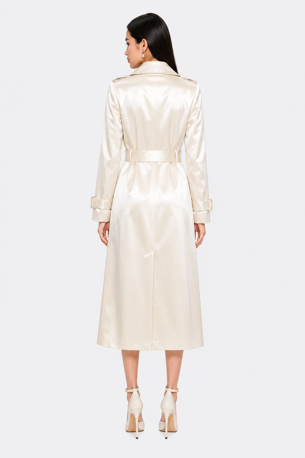 A woman viewed from the back wearing a long cream satin trench coat with a matching waist belt, paired with white high heels, against a light gray studio background.