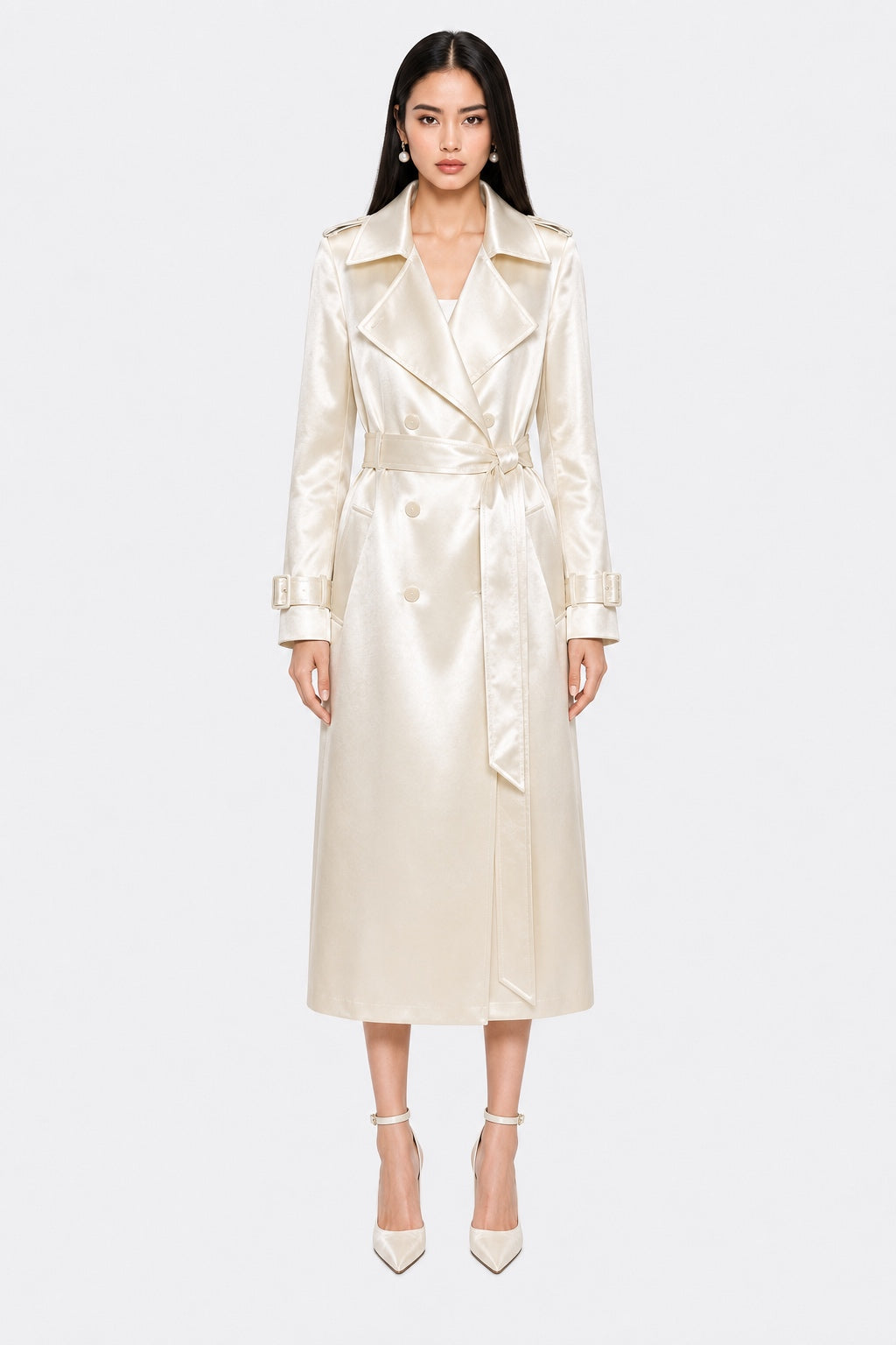 A model wearing a long, cream-colored satin trench coat with a matching waist tie belt, paired with matching pointed-toe heels, set against a plain light gray background.