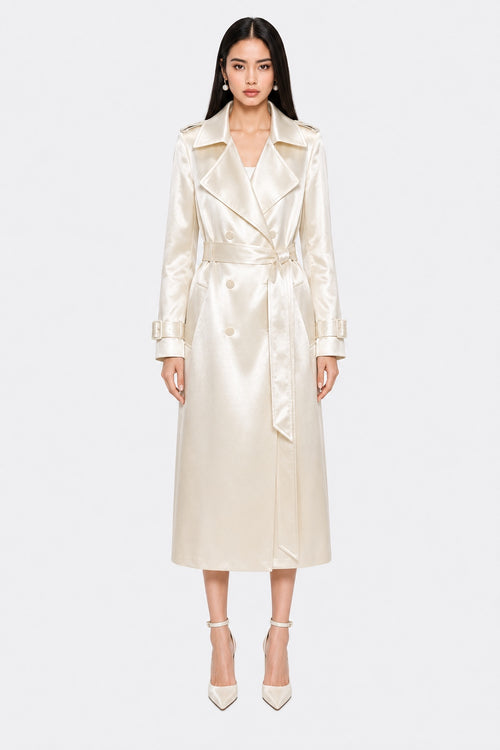 A model wearing a long, cream-colored satin trench coat with a matching waist tie belt, paired with matching pointed-toe heels, set against a plain light gray background.