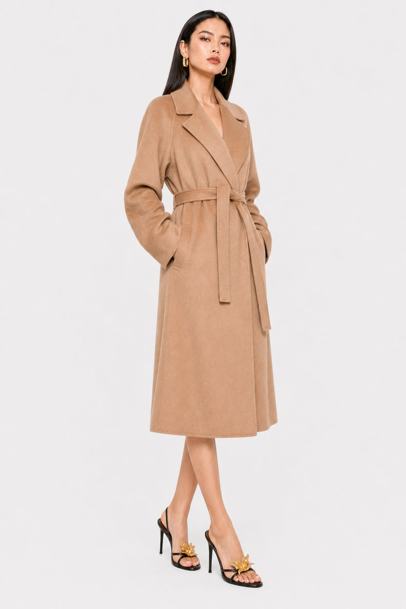 A woman modeling a mid-length camel brown wrap coat with a self-tie belt, paired with black high heels, against a plain light gray studio background.