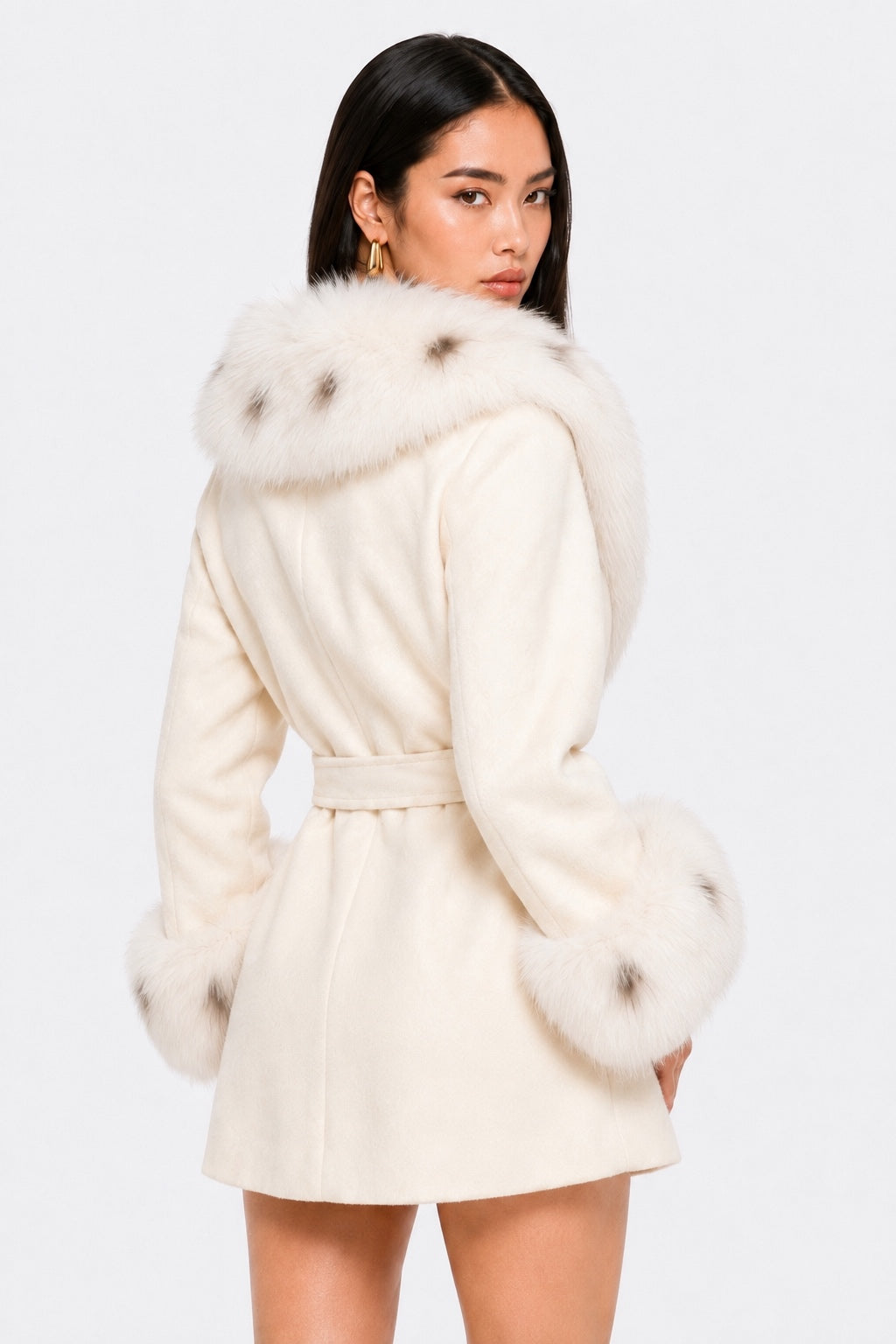 Back three-quarter view of a model wearing an ivory belted wool coat with a full fox fur collar and fluffy fox fur cuffs