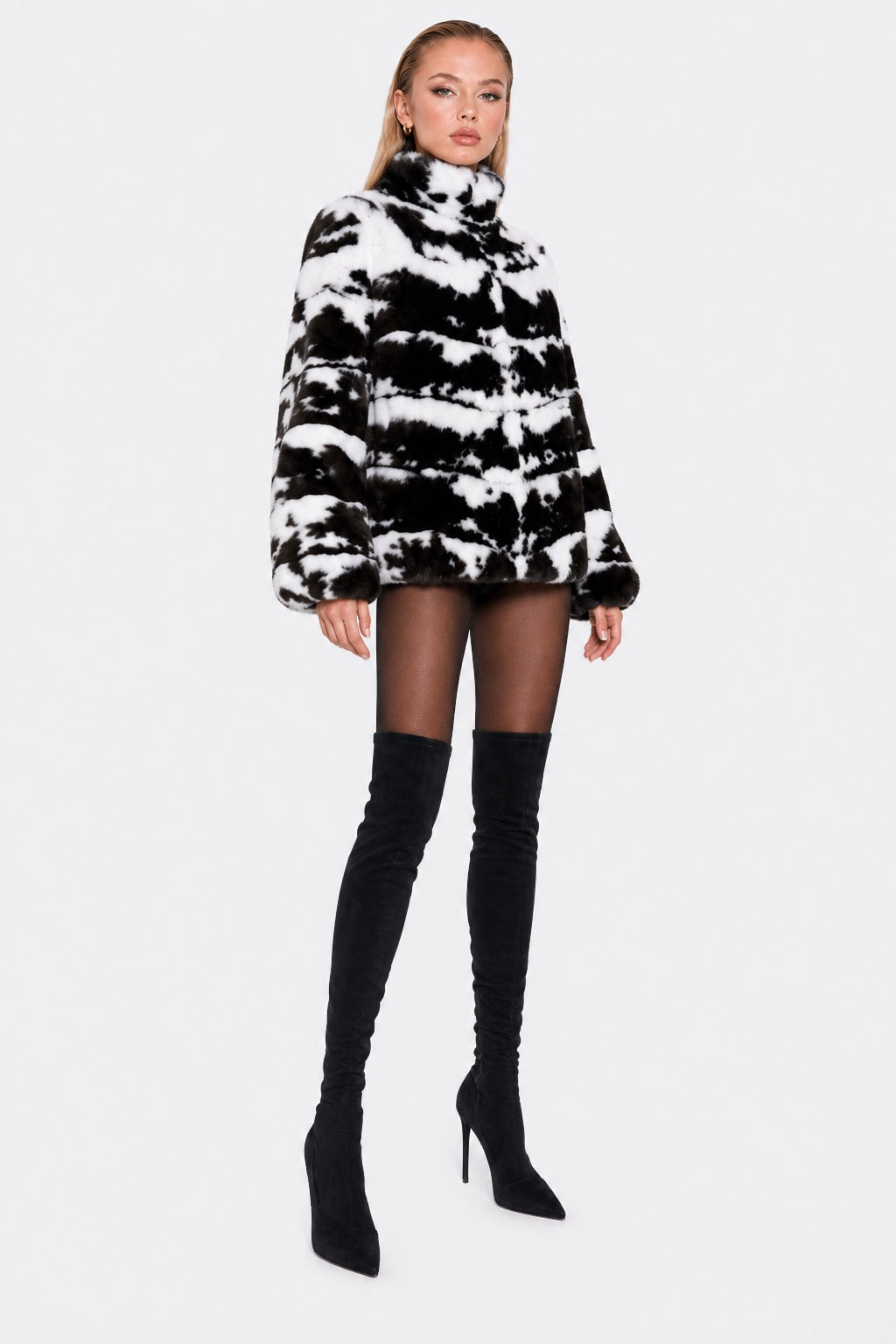 A model wearing a black and white tie-dye faux fur turtleneck jacket, sheer black tights, and black over-the-knee boots, standing against a light gray studio background.
