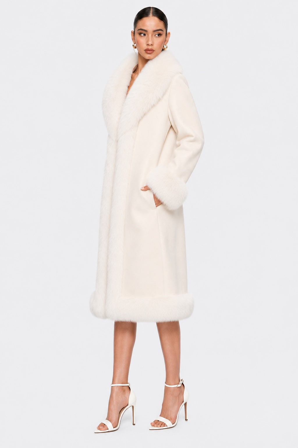 A model wearing a long cream natural fur coat with a wide collar and matching cuffs, paired with white high-heeled sandals, against a light gray studio background.