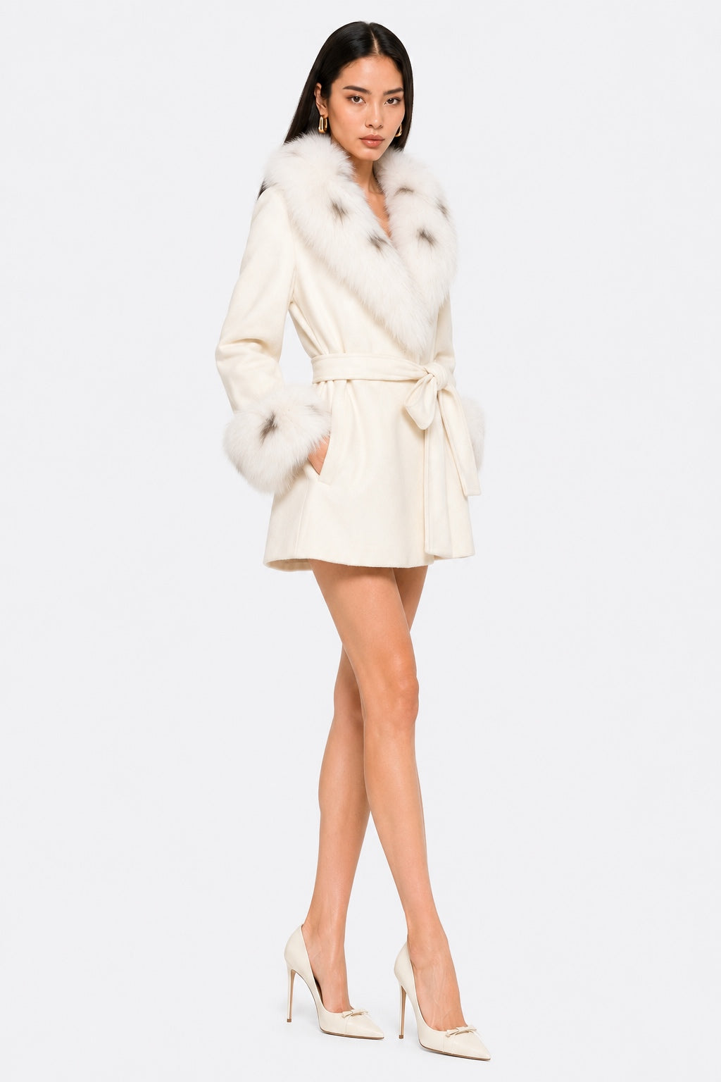 Model wearing an ivory 100% wool belted coat with a large plush fox fur collar and matching fox fur cuffs, shown in a three-quarter front pose with both hands in the pockets