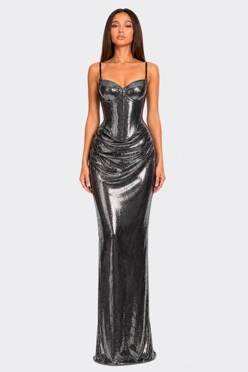 A woman modeling a floor-length, metallic silver sequined gown with spaghetti straps, a sweetheart neckline, and a ruched corset-style bodice against a light gray studio background.