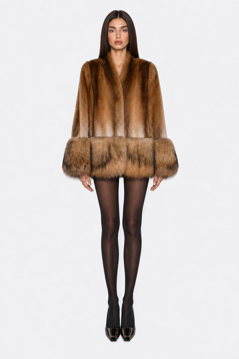 A model wearing a brown fur coat with a wide collar and voluminous cuffs, paired with black tights and black pointed-toe heels, shot against a solid light gray background.
