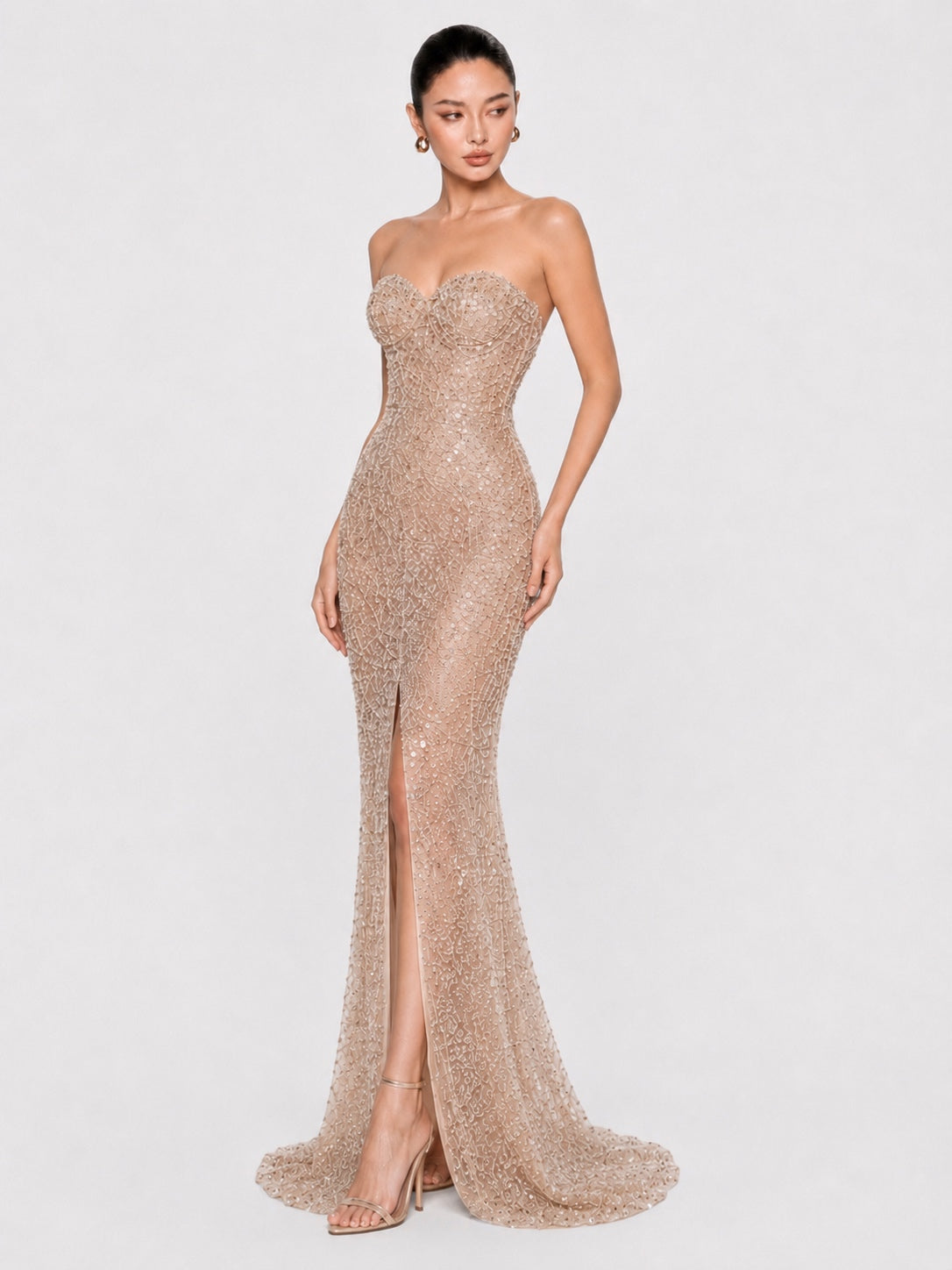 A woman modeling a strapless, floor-length champagne beige gown covered in shimmering sequins and beads, featuring a sweetheart neckline, fitted silhouette, thigh-high slit, and a slight train against a light gray studio background.