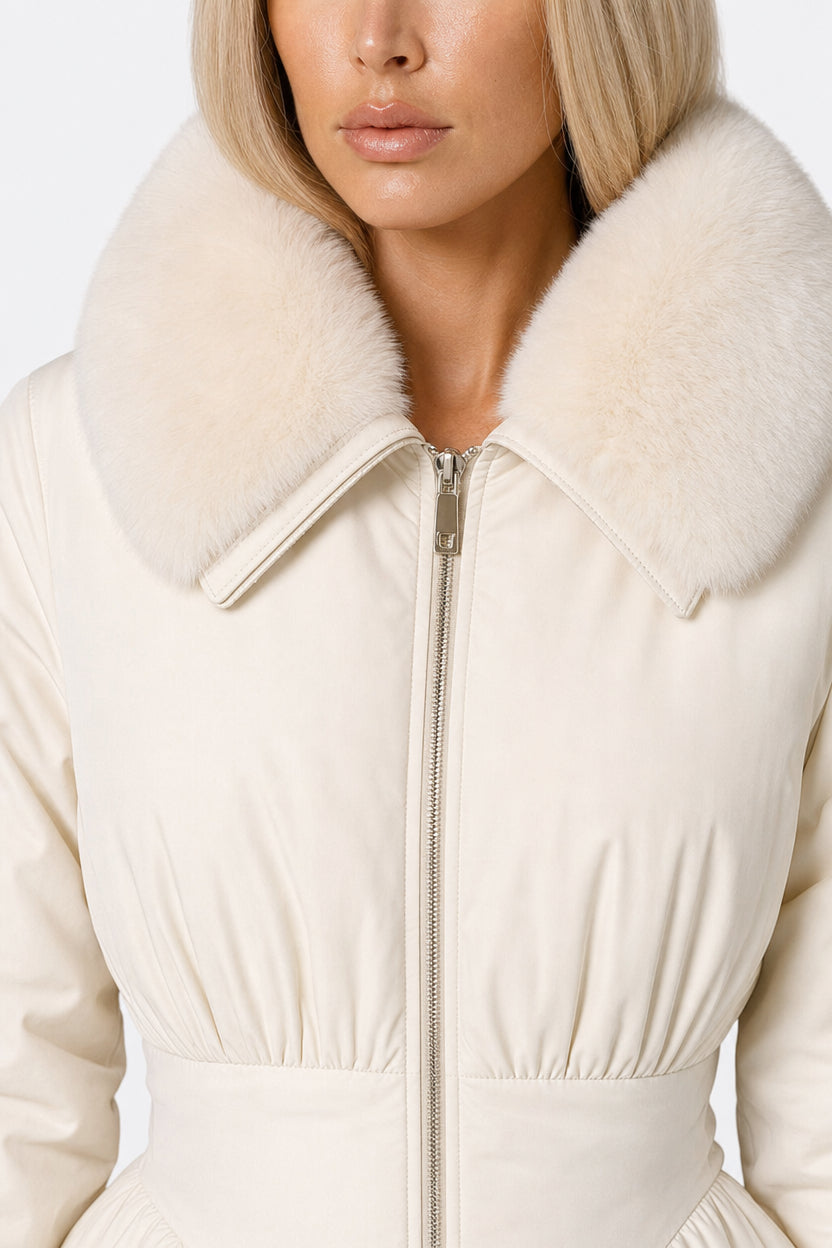A close-up of a model wearing a cream-colored puffer jacket with a large, fluffy faux-fur collar and a front zipper.