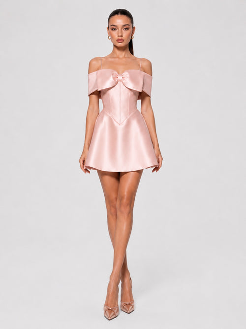 A woman modeling a light pink off-the-shoulder satin mini dress with a large bow detail at the bust, paired with clear high-heeled sandals, against a light gray studio background.