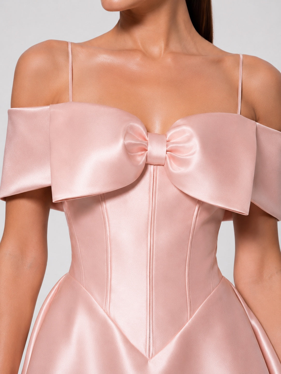 A close-up of a model wearing a light pink satin dress with a large bow at the bust, thin spaghetti straps, and a structured corset-style bodice.