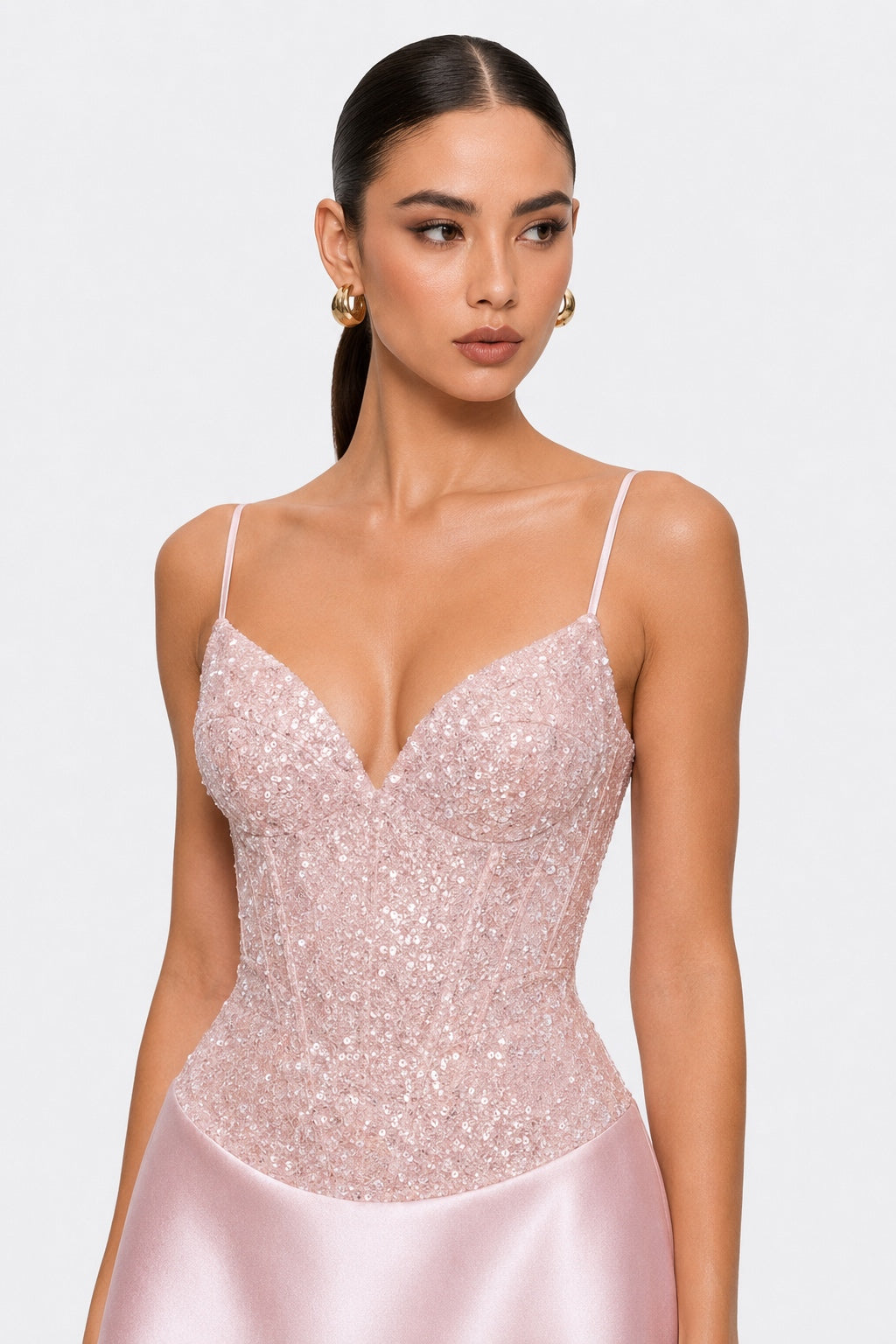 A woman modeling a light pink sequined corset-style dress with thin spaghetti straps and a satin skirt against a plain light gray studio background.