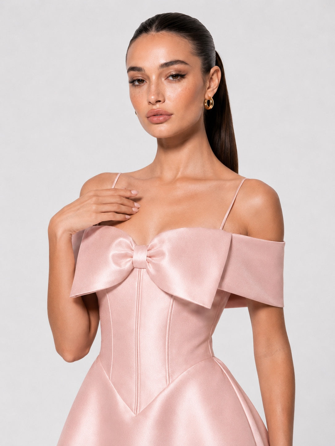 A woman modeling a pale blush pink satin off-the-shoulder dress with a large bow at the bust and thin spaghetti straps, photographed against a light gray studio background.