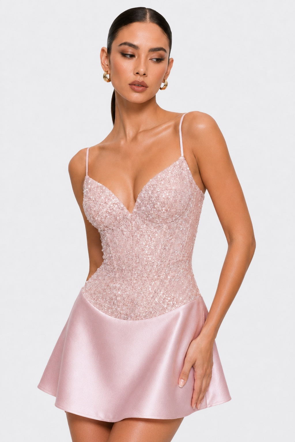 A woman modeling a light pink spaghetti-strap mini dress with a sequined corset-style bodice and a satin flared skirt, photographed against a plain light gray studio background.