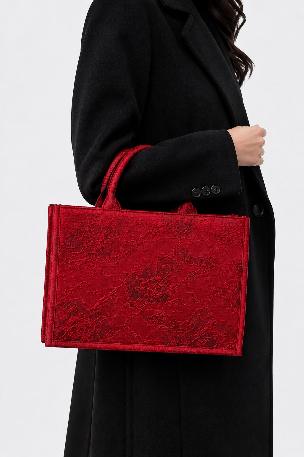 A woman in a black coat carries a structured red tote bag with a textured, slightly glossy surface and matching red handles against a light gray background.