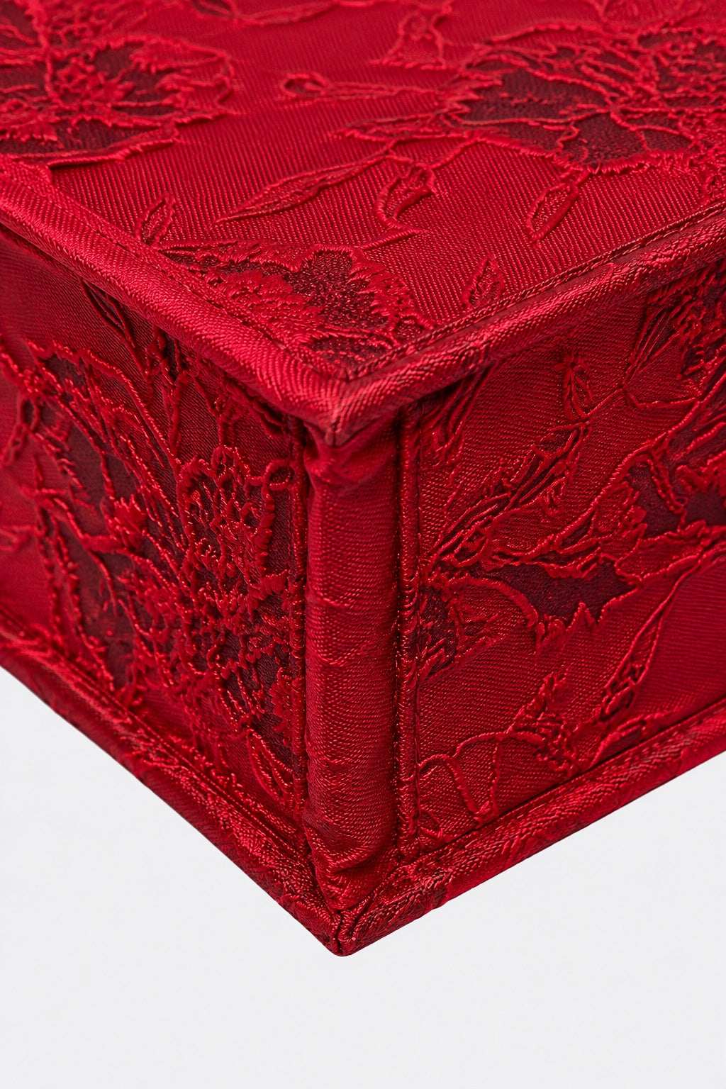 A close-up of a red embroidered fabric box corner, showing detailed floral lace embroidery and satin-like texture against a light gray background.