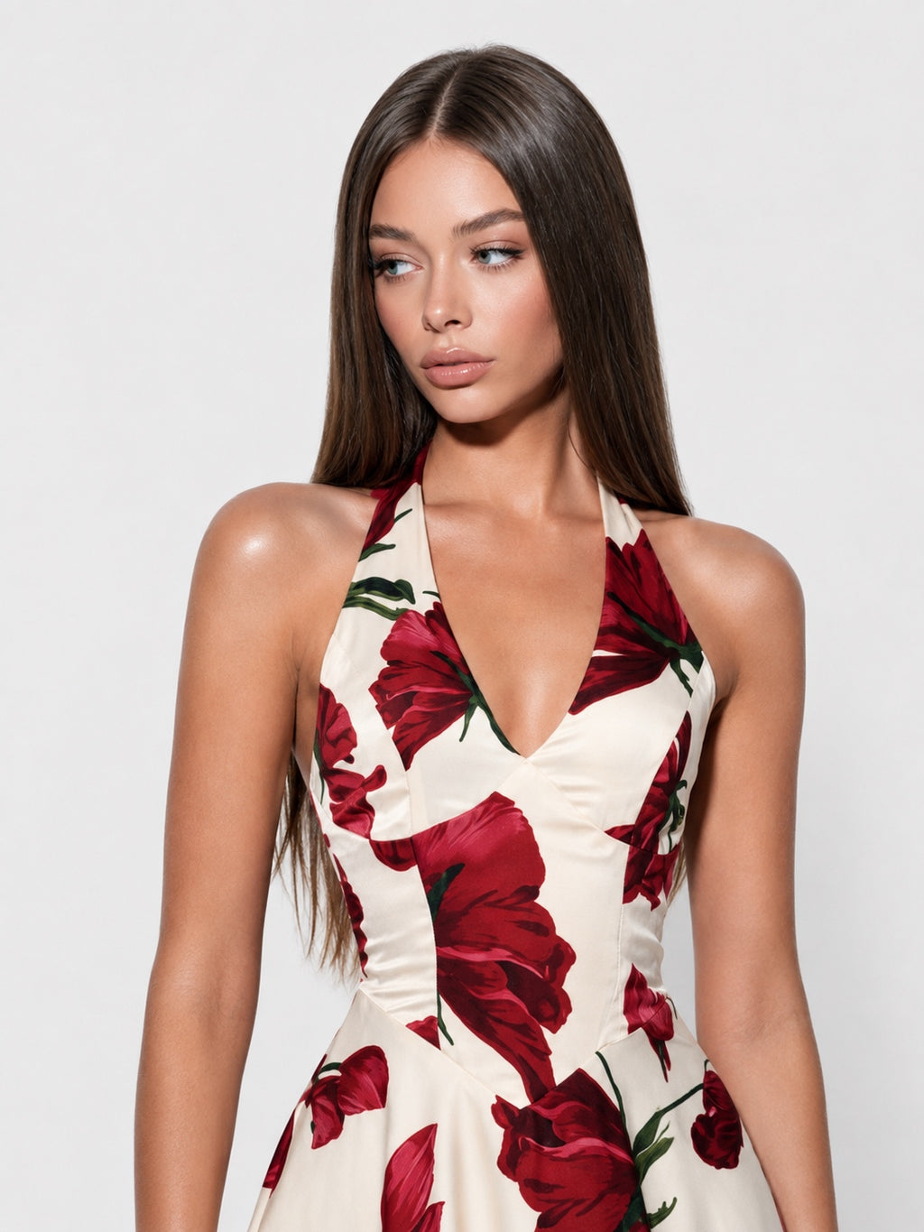A woman modeling a halter-neck dress with a deep V-neckline, featuring a red and green floral print on a cream-colored background, set against a plain light gray studio backdrop.