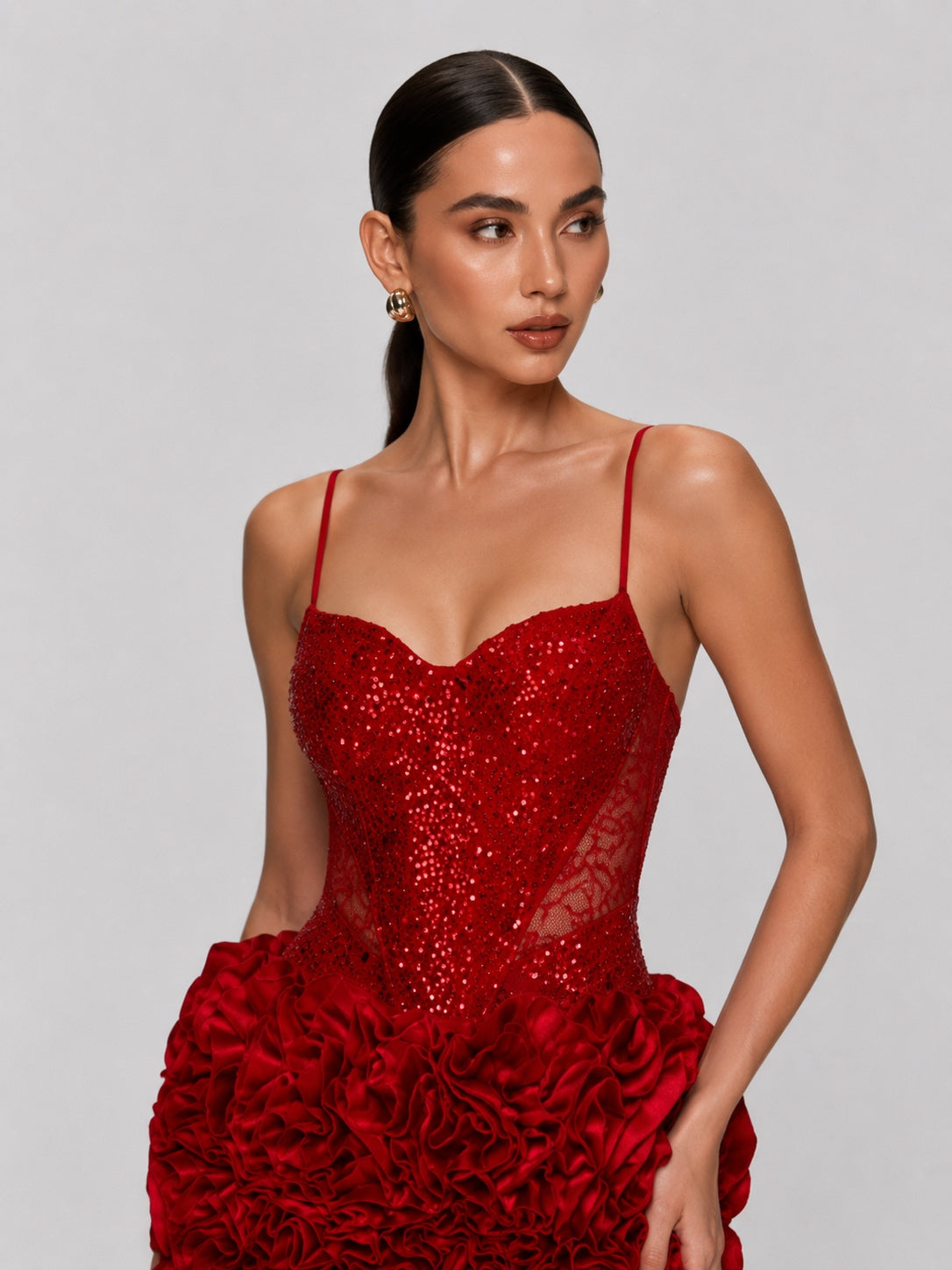 A woman modeling a red evening gown with thin spaghetti straps, a sweetheart neckline, a sequined corset-style bodice, and a voluminous ruffled skirt, photographed against a solid gray studio background.