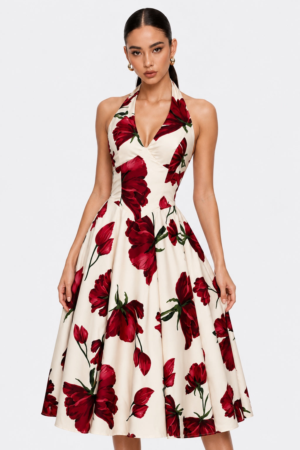 A woman modeling a sleeveless halter-neck midi dress with a deep V-neckline and a flared skirt, featuring a bold red rose floral print on an off-white background, set against a solid light gray studio backdrop.
