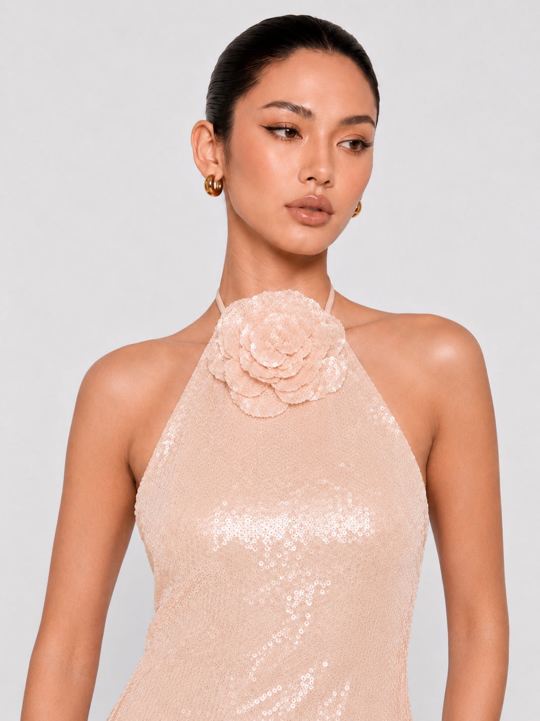 A woman wearing a pale blush pink sequined halter dress with a large fabric flower applique at the neckline, photographed against a light gray studio background.