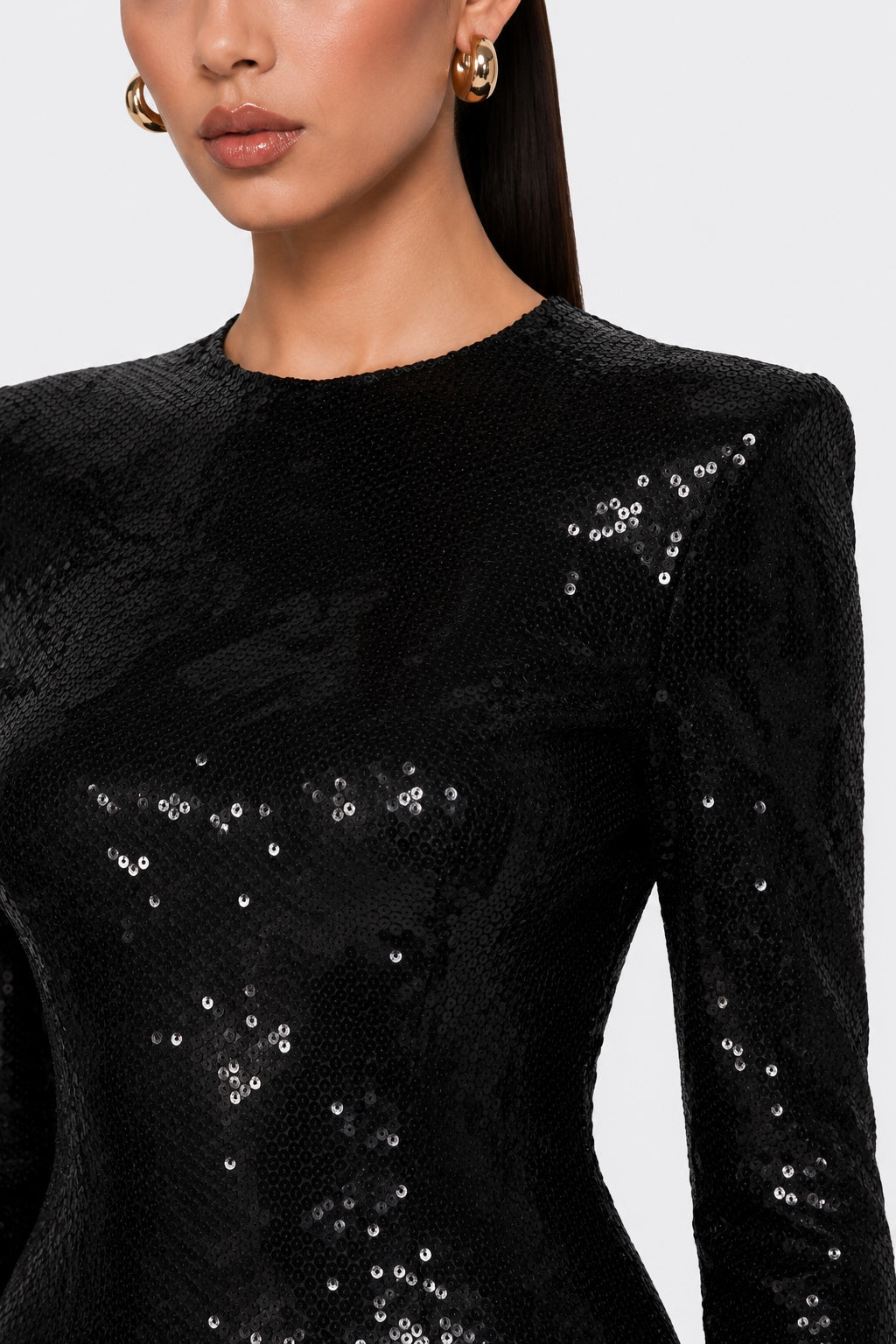 A close-up of a model wearing a black sequined long-sleeve top with structured shoulders and gold hoop earrings against a light gray background.