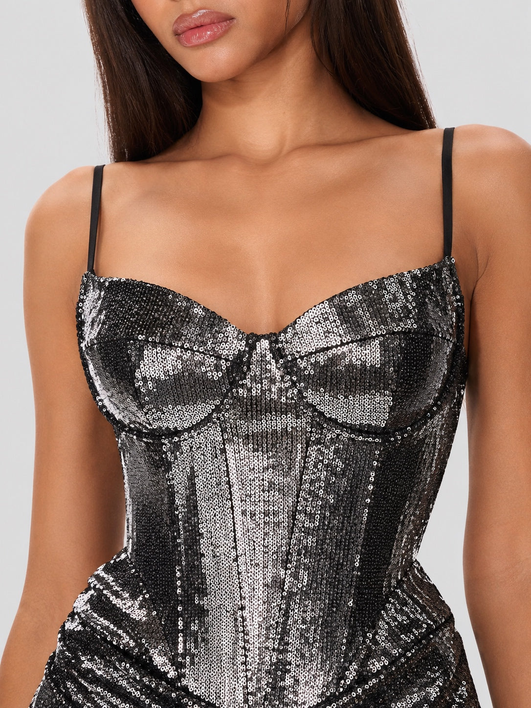 A close-up of a model wearing a black and silver sequined corset-style camisole dress with thin straps against a light gray background.