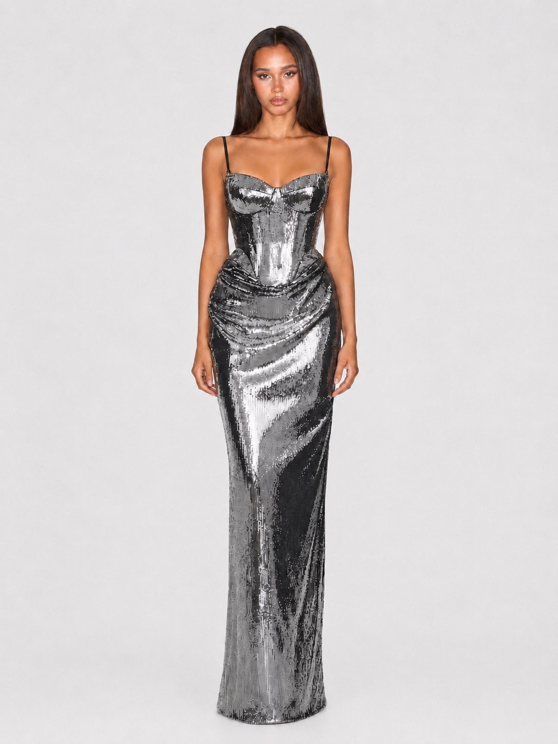 A woman modeling a floor-length metallic silver sequin gown with spaghetti straps and a sweetheart neckline against a light gray studio background.