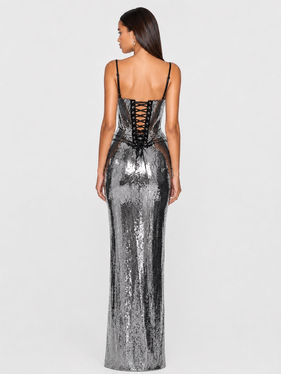 A woman shown from the back wearing a floor-length silver sequin gown with thin black straps and a lace-up corset-style back, photographed against a light gray studio background.
