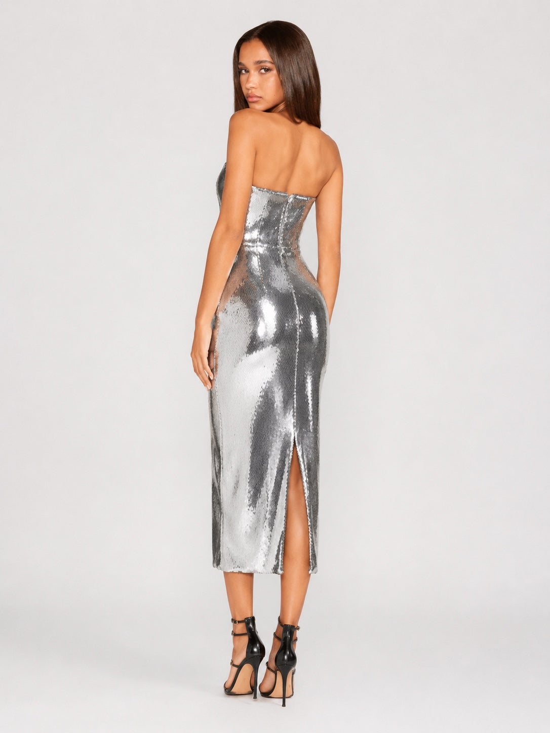 A woman modeling a strapless metallic silver midi dress with a sweetheart neckline, fitted silhouette, and front slit, shown from the back against a light gray studio background.